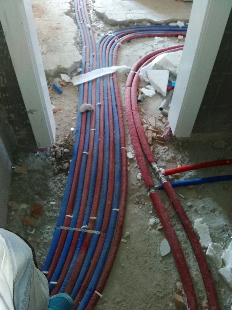 Egy építkezés vagy teljes felújítás alatt álló lakás padlója látható, egy ajtónyíláson keresztül fényképezve. A nyers, feltört betonpadlón kék és piros szigeteléssel ellátott fűtéscsövek kötegei futnak ívesen a földön. Jobb oldalon további piros és kék csövek csatlakoznak egymáshoz T-idomokkal. A padlón mindenütt építési törmelék, vakolatdarabok és fehér hungarocell hulladék hever.