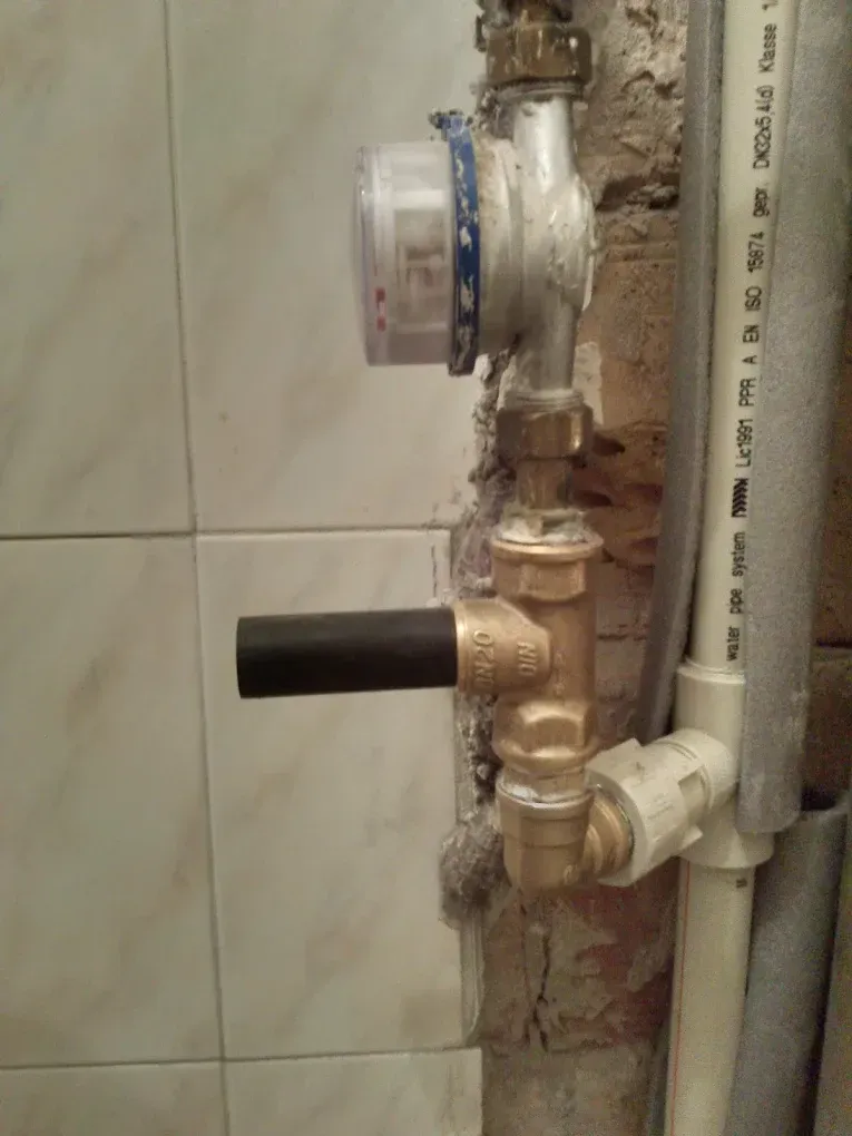 Közeli felvétel egy falban lévő, szerelés alatt álló vízvezeték-szakaszról. Felül egy analóg vízóra látható, amihez alul egy sárgaréz T-idom csatlakozik, amelyből balra egy fekete, henger alakú műanyag toldat áll ki. Ehhez a réz idomhoz alulról egy szürke szigeteléssel ellátott, feliratos fehér műanyag (PPR) vízvezeték csatlakozik egy könyökidommal.