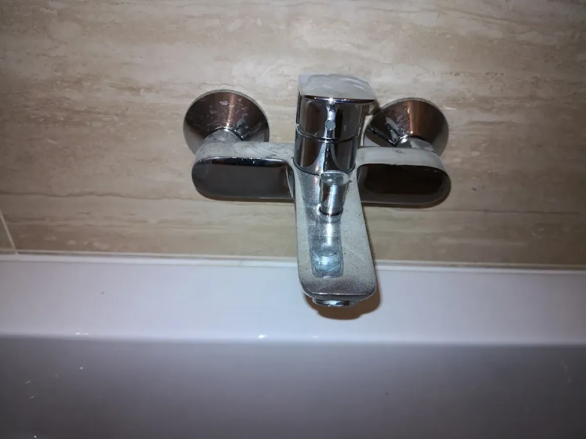 Közeli felvétel egy krómozott, fali kádcsaptelepről. A csap egykaros, felül található a keverőkar, a rövid, lefelé néző kifolyócső tetején pedig egy apró zuhanyváltó gomb van. A csaptelep mögött világos, bézs színű, márványos mintázatú csempe látható, alatta pedig a fehér fürdőkád pereme húzódik.