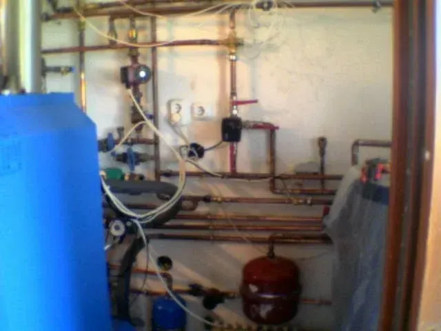 Egy kissé elmosódott kép egy kazánház vagy gépészeti helyiség faláról, amely tele van bonyolultan futó, függőleges és vízszintes vörösréz fűtéscsövekkel. A csövekre két fekete-piros keringető szivattyú és több piros karos golyóscsap van szerelve. A kép bal oldalát egy nagy kék tartály (bojler vagy puffer) uralja, lent a padlón pedig egy piros tágulási tartály ül. A falon konnektorok és lazán lógó elektromos vezetékek láthatók.