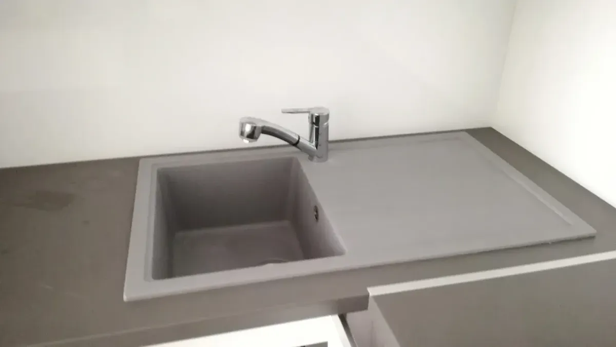 Egy modern, letisztult konyhai mosogatótálca látható, amely matt sötétszürke, gránithatású anyagból készült, egy csepegtetőtálcás résszel a jobb oldalon. A mosogatóba egy elegáns, egykaros krómozott csaptelep van beszerelve. A pult is sötétszürke színű, a hátfal és a pult alatti szekrényrészek pedig fehérek.