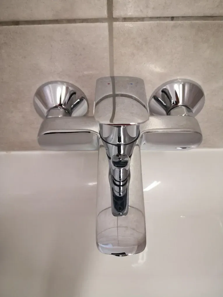 Felülnézeti, közeli kép egy modern, fényesre krómozott fali csaptelepről, amely egy fehér mosdókagyló vagy kád fölé van felszerelve. A csaptelep egykaros, a lapos fogantyú tetején apró piros és kék pöttyök jelzik a meleg és hideg vizet. A háttérben világos, semleges színű csempe látható.