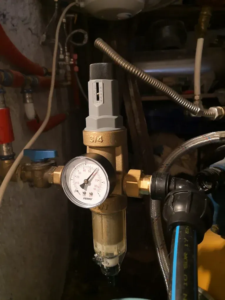 Sötét, gépészeti térben készült közeli felvétel egy sárgaréz nyomáscsökkentő szelepről és vízszűrőről. A szerelvény bal oldalán egy kerek, FERRO márkájú nyomásmérő óra (manométer) látható, felül pedig egy szürke-fekete műanyag tekerőgomb egy mércével. A rendszerhez alulról egy kék csíkos vastag fekete KPE cső, jobbról és felülről pedig fém flexibilis csövek és többrétegű műanyag csövek csatlakoznak, kék és piros elzárócsapokkal a háttérben.