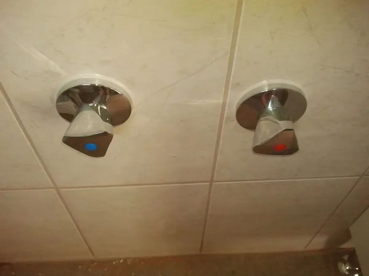 Két krómozott, falba süllyesztett csaptelep-fogantyú (elzárószelep) látható, világos bézs, enyhén márványos mintázatú csempén. A fogantyúk háromszög alakúak, lekerekített sarkokkal. A bal oldaliban egy kis kék pötty, a jobb oldaliban pedig egy piros pötty található, melyek a hideg, illetve a meleg vizet jelzik.