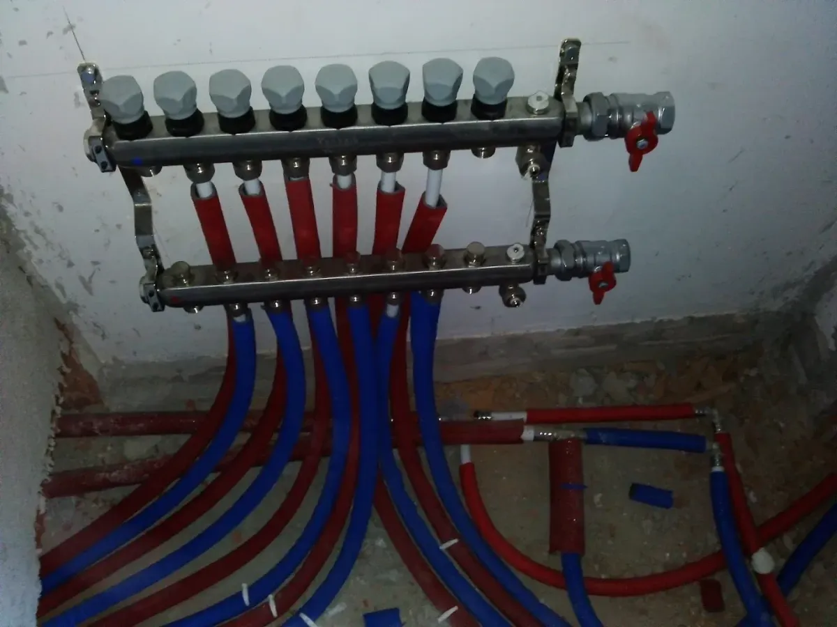Egy padlófűtési rendszer vagy vízhálózat rozsdamentes acél osztó-gyűjtője (elosztóblokkja) látható a falra szerelve. A felső sávon szürke kupakos áramlásmérők vagy szelepek, az alsón pedig további csatlakozók találhatók. Mindkét rúd végén egy-egy piros karos golyóscsap van. Az elosztóból lefelé kék és piros szigetelőcsőbe (gégecsőbe) húzott vezetékek futnak a nyers betonpadló felé.