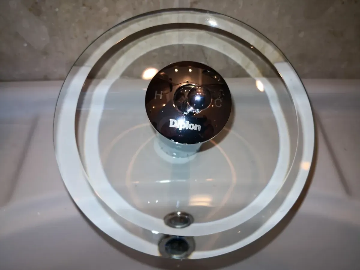 Felülnézeti kép egy különleges, modern mosdócsaptelepről, amely egy kerek, átlátszó üvegtányérból áll (vízesés-kifolyó), középen egy krómozott fémhengerrel. A fémen a "Diplon" márkanév, valamint a hideg-meleg vizet jelző C és H betűk olvashatók. A csaptelep egy fehér kerámia mosdókagylóra van szerelve, a háttérben világos színű, márványos csempe látszik.