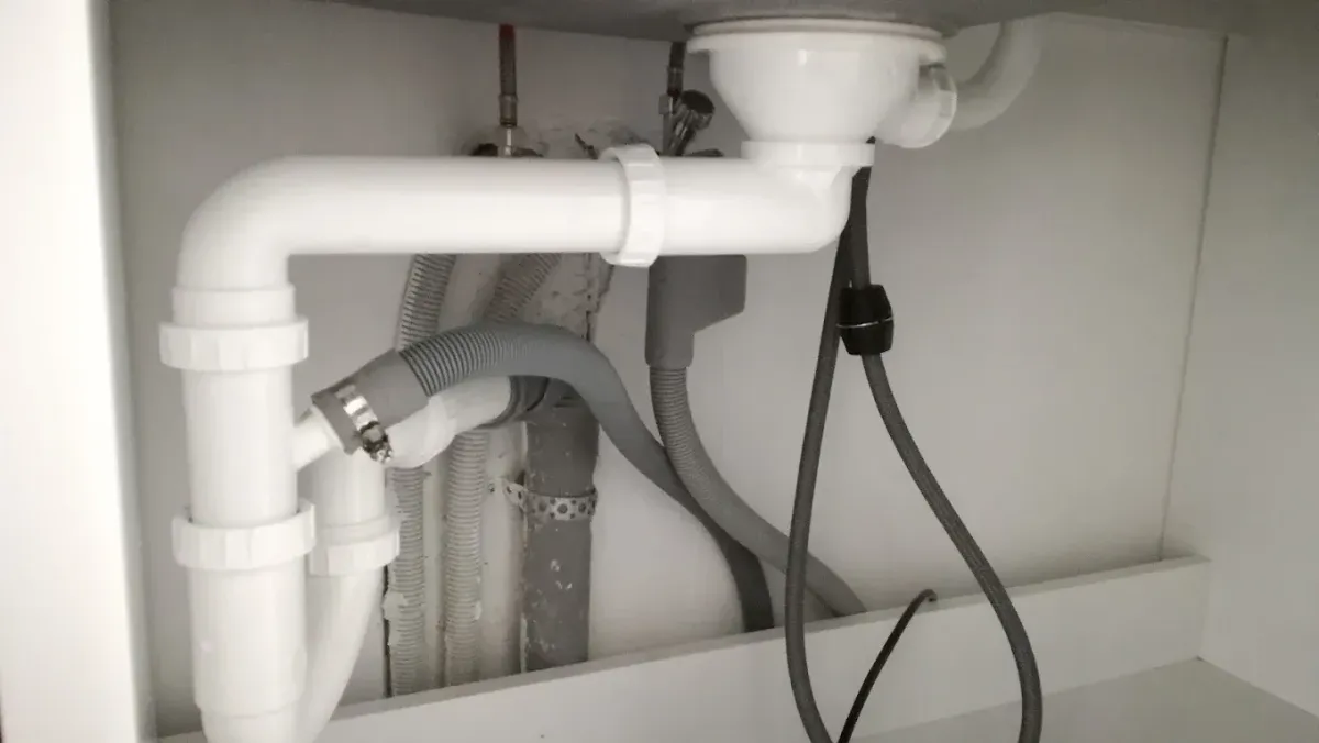 Mosogató alatti szerelvények egy szekrényben. Középpontban a fehér műanyag U-szifon. Mögötte szürke fali lefolyócső, abba bekötött bordázott szürke mosogatógép cső bilinccsel, valamint sarokszelepek és flexibilis vízcsövek. Jobb oldalon egy fekete kábel és egy súly látható lógva.