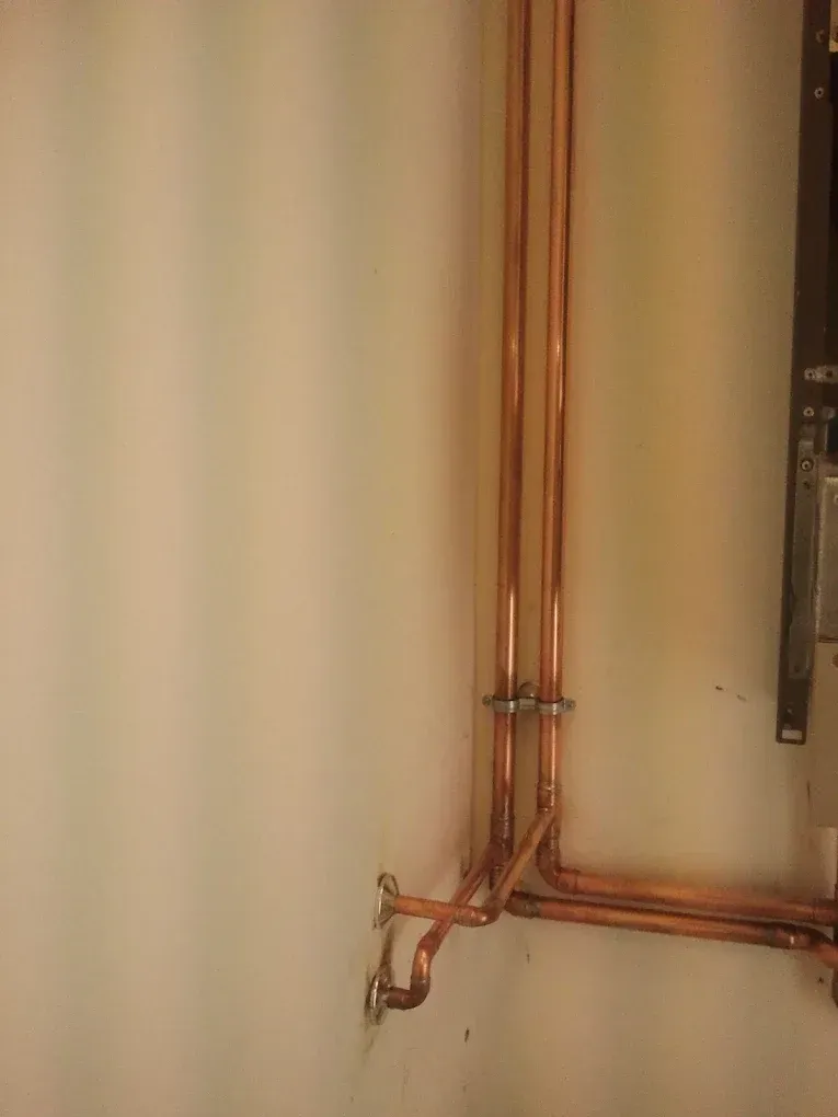Két párhuzamosan futó, fénylő vörösréz fűtéscső látható egy fehérre festett fal sarkában. A csövek függőlegesen haladnak lefelé, majd a padlóhoz közel 90 fokban jobbra kanyarodnak. A csöveket fém bilincsek rögzítik a falhoz, a forrasztási pontoknál pedig ezüstös elszíneződések látszanak a rézen.