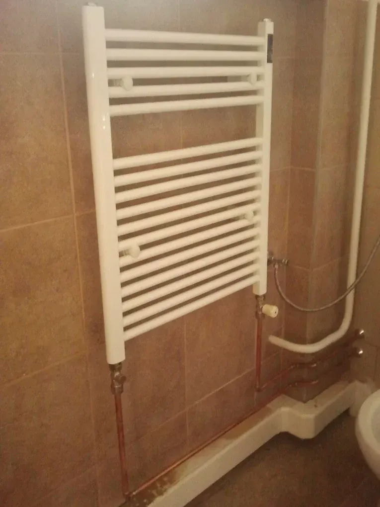Egy fehér színű, csöves törölközőszárító radiátor van felszerelve egy barna, márványmintás csempével burkolott falra. A radiátorhoz alulról vékony rézcsövek csatlakoznak, amelyek a falon kívül haladnak a fehér padlószegély felett. Jobb oldalon, a padló közelében egy fehér szaniter (vécé vagy bidé) egy kis része lóg a képbe.
