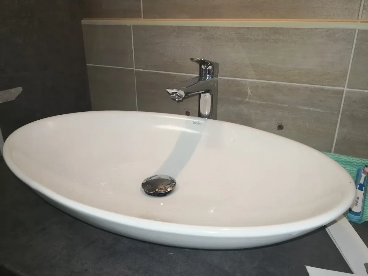 Egy elegáns, ovális alakú, pultra ültetett, modern fehér kerámia mosdókagyló ("baliv" felirattal) látható egy sötétszürke pulton. A mosdóhoz egy szögletes vonalvezetésű, krómozott fali csaptelep tartozik. A mosdóban még vízcseppek láthatók. A hátteret vízszintes, téglalap alakú, bézs/szürkés árnyalatú csempék alkotják, amelyekben fúrt lyukak (esetleg korábbi szerelvények nyomai) látszanak. Jobb oldalon egy fogkefetartó széle és egy tubus fogkrém lóg a képbe.