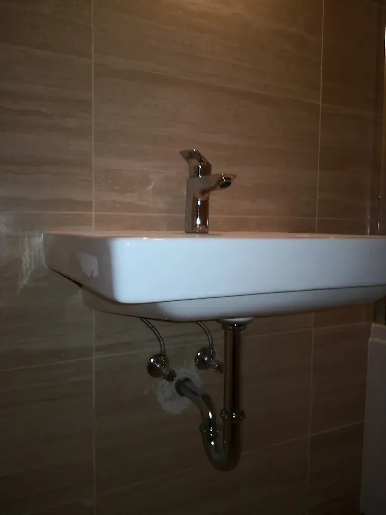 Egy modern, szögletes formájú, fehér fali mosdókagyló látható profilból fényképezve. A mosdón egy egykaros krómozott csaptelep van. A mosdó alatt jól látható a krómozott szifon (lefolyócső) és a falból kiálló sarokszelepek, amelyekhez flexibilis csövek csatlakoznak. A fali lefolyókiállás körül némi fehér tömítőanyag vagy gipsz látszik. A háttérben lévő falat vízszintesen mintázott, bézs színű csempe fedi, a mosdó bal elülső peremén pedig egy kis darab fehér ragasztószalag vagy fólia maradványa tapad.