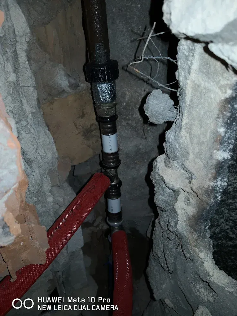 Közeli felvétel egy vastag, bontott falban lévő csővezetékről. Egy felülről érkező, régi fekete fémcsövet fekete présidomokkal toldottak össze egy modern, fehér ötrétegű csővel. A csőhálózathoz egy T-idommal csatlakozik egy piros gégecsőbe (szigetelésbe) húzott ág is, amely balra lefelé tart a törmelékes üregben. A kép alján Huawei vízjel található.