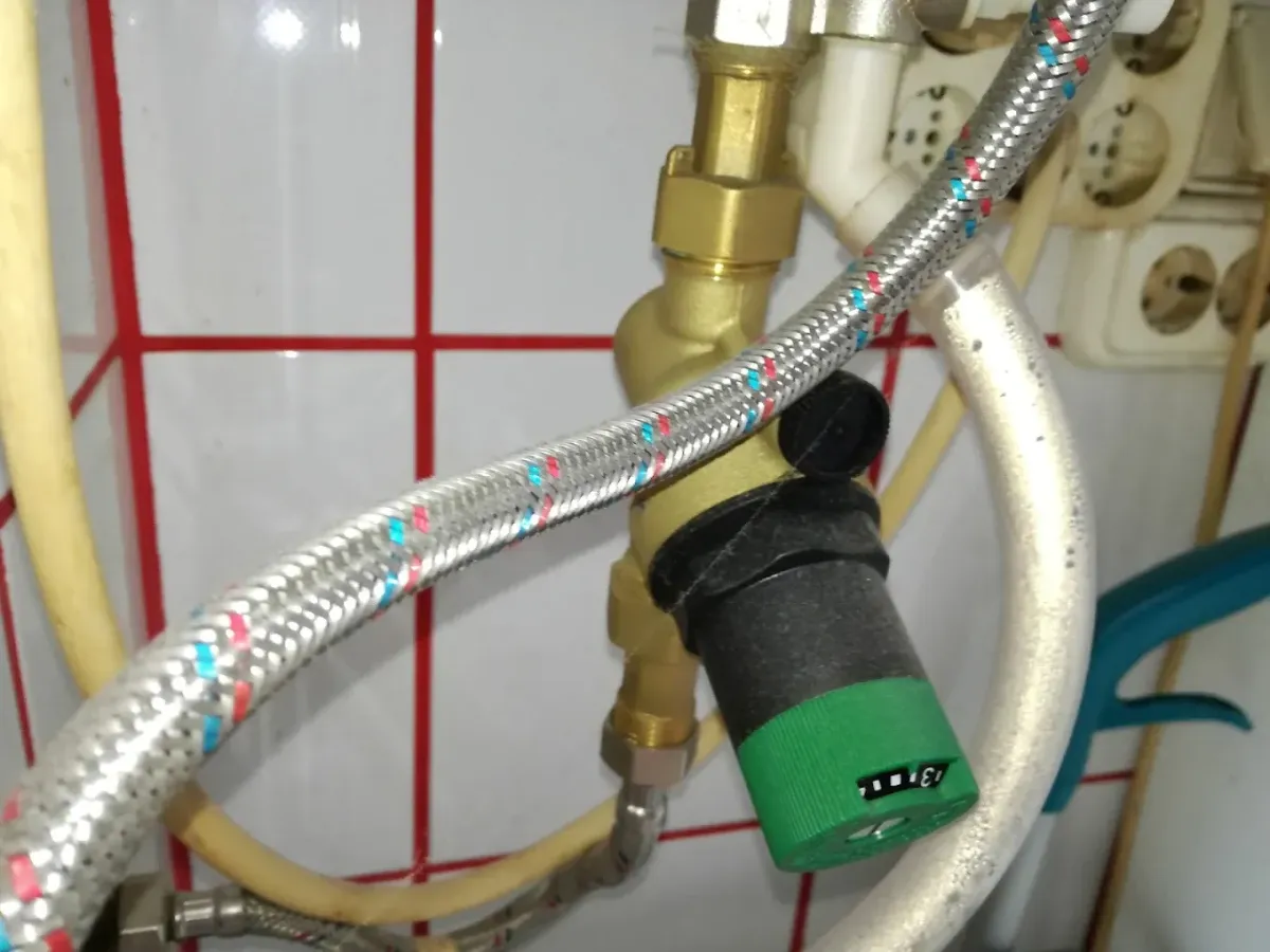 Egy sárgaréz vízvezeték-szerelvény, valószínűleg egy nyomáscsökkentő szelep látható közelről, amelynek az alján egy nagy, zöld színű műanyag tekerőgomb van, számskálával. A kép előterében egy fémhálós, piros-kék jelölésű flexibilis cső fut keresztbe. A háttérben fehér csempe piros fugával, valamint egy falra szerelt, kissé elszíneződött elektromos elosztó dugalj látható.