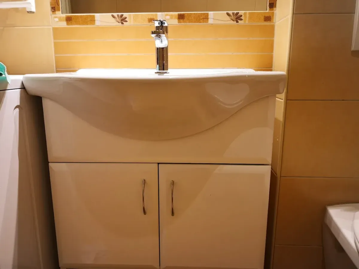 Egy fürdőszobai mosdószekrény látható. A szekrény alja fényes fehér, két ajtóval és fém fogantyúkkal, tetején pedig egy elegánsan ívelt, fehér kerámia mosdókagyló ül, rajta egy krómozott csapteleppel. A mosdó mögötti falat sárgás-narancssárgás, meleg tónusú csempe borítja, amely felett egy virágmintás díszítőcsík és egy tükör alja látható.