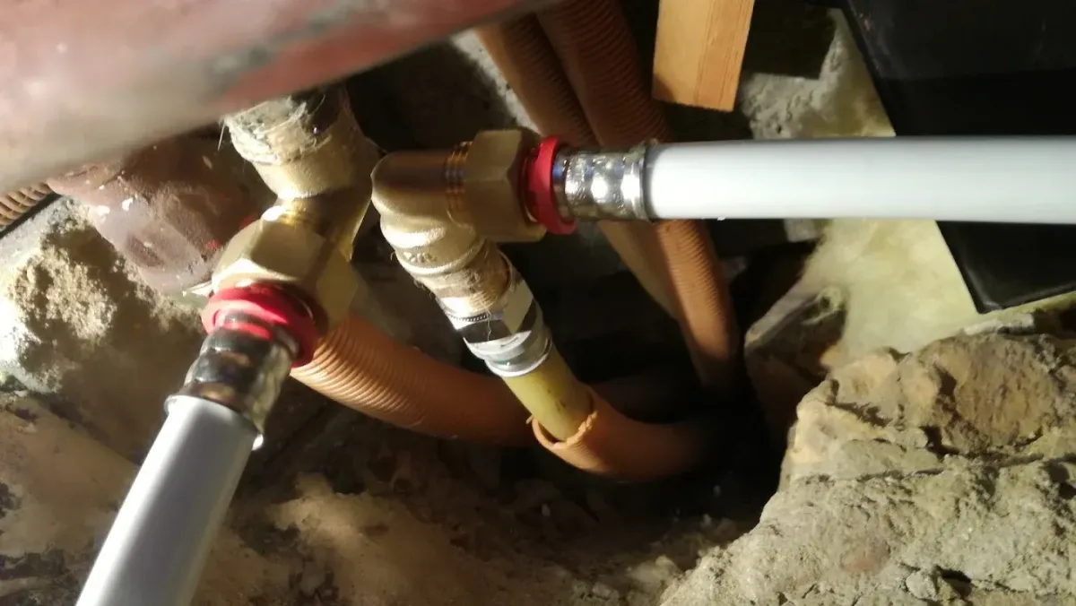 Szűk, bontott fal- vagy padlórészletben futó vízvezetékek közelképe. Két fehér színű, modern ötrétegű cső csatlakozik sárgaréz idomokkal (könyök és T-idom) és piros présgyűrűkkel egy régebbi, fém csőhálózathoz. A csövek körül narancssárga bordázott védőcsövek (gégecsövek) futnak be a fal vagy a födém mélyébe, a háttérben törmelék és kőzetgyapot szigetelés darabjai látszanak.