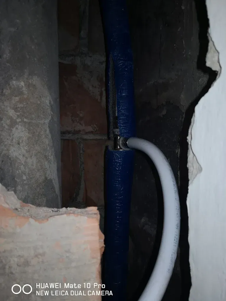 Sötét felvétel egy bontott falban futó vízvezetékről. Egy függőlegesen futó, vastag kék szigeteléssel bevont csőből egy T-idom segítségével egy fehér ötrétegű műanyag cső ágazik le jobbra, majd ívesen lefelé hajlik. A csövek körül nyers tégla, beton és a kibontott fal durva szélei látszanak. A kép alján "HUAWEI Mate 10 Pro" vízjel olvasható.