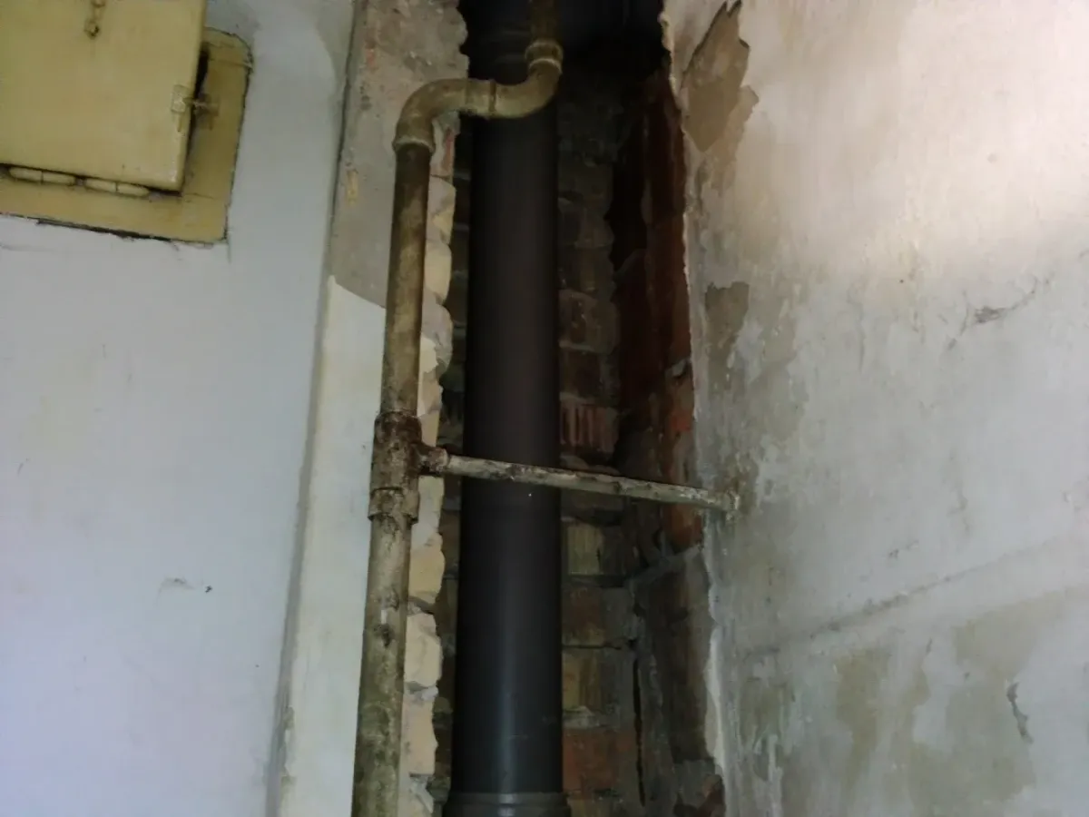 Egy falba vájt bontott nyílás (horony) látható, amelyben egy vastag, sötétszürke műanyag lefolyócső (PVC) fut függőlegesen a nyers téglafal előtt. Előtte egy vékonyabb, régi, rozsdás fémcső is halad, amelyből egy vízszintes ág keresztezi a vastag lefolyót. A bal oldali ép, fehérre festett falon egy kisméretű, krémszínű szerelőajtó is helyet kapott.