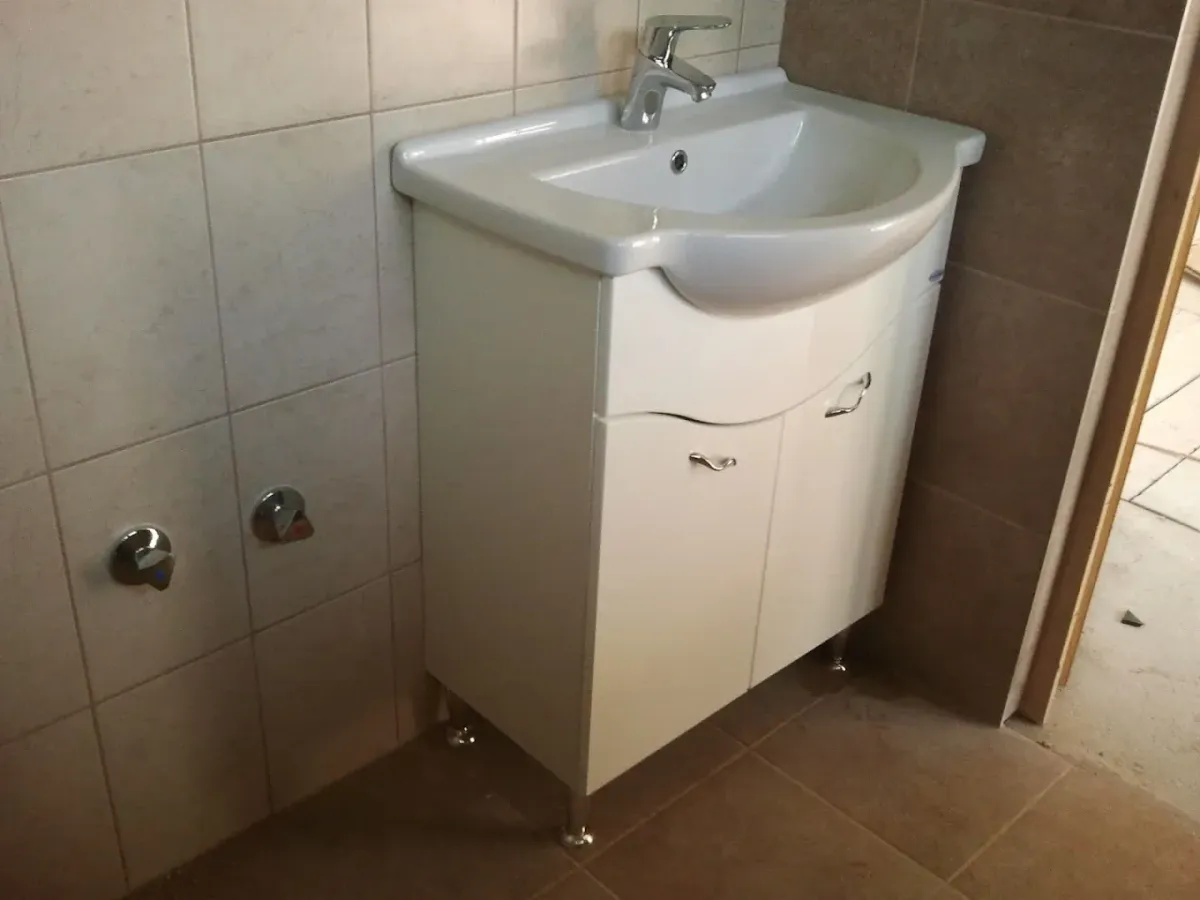 Egy fürdőszobai, fehér mosdószekrény látható beépítés alatt vagy után. A szekrény fényes, íves ajtókkal és króm fogantyúkkal rendelkezik, ezüstös fém lábakon áll a barnás padlócsempén. Tetején egy fehér kerámia mosdókagyló és egy egykaros csaptelep van. A szekrénytől balra, a világos falicsempén két krómozott, háromszögletű sarokszelep (elzárócsap) áll ki a falból. Jobbra egy nyitott ajtónyílás látszik.