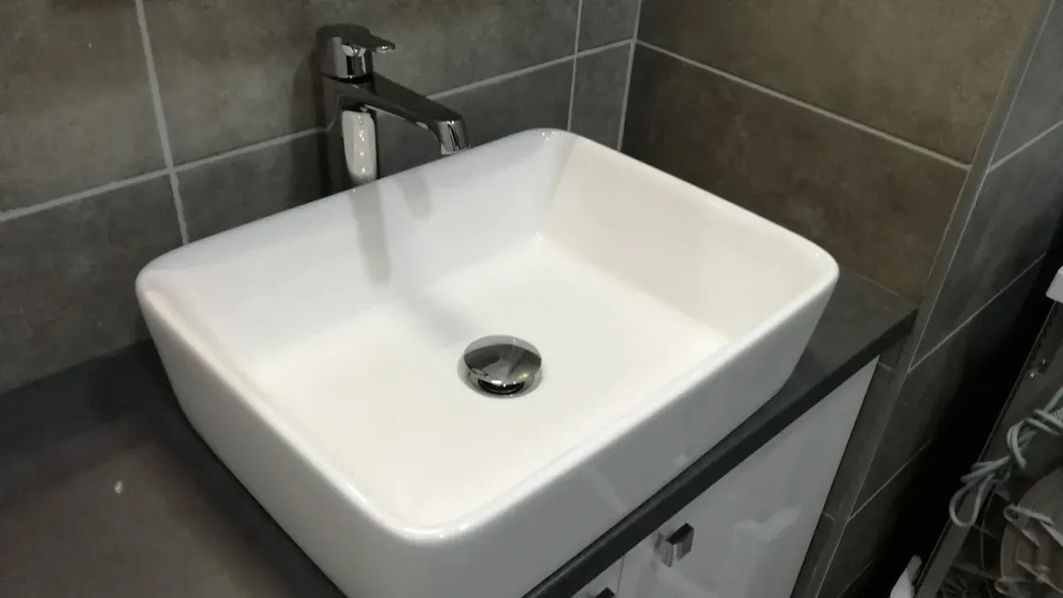 Egy modern fürdőszobai mosdó látható. A pultra ültetett, szögletes, mégis finoman lekerekített sarkú fehér kerámia mosdókagyló egy sötétszürke pulton áll, amely alatt egy fehér, fényes felületű, króm fogantyús szekrény található. A mosdóhoz egy elegáns, magasított egykaros krómozott csaptelep tartozik. A hátteret nagyméretű, barna és szürkésbarna árnyalatú, kőhatású csempék alkotják.