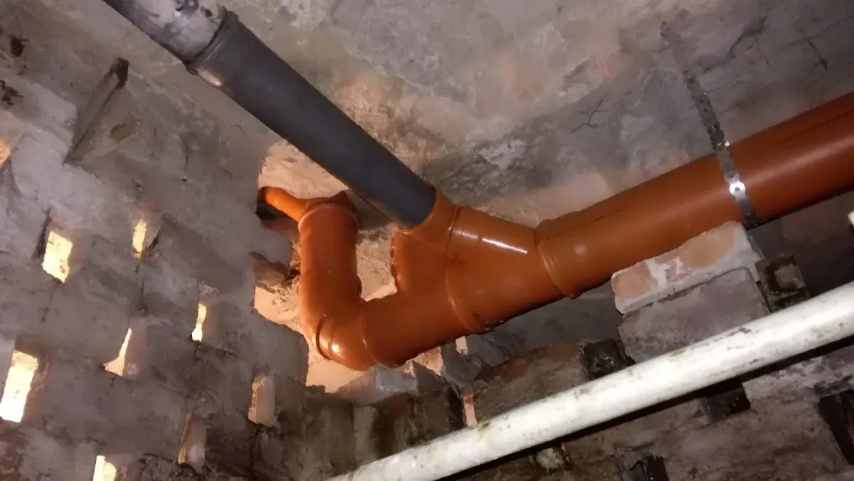 Alulról felfelé fényképezett jelenet egy pincében vagy alagsorban, ahol lefolyócsövek haladnak át a födémen. Egy vízszintesen futó narancssárga PVC csatornacső egy könyökidommal csatlakozik egy vastag szürke csőhöz, amely eltűnik a mennyezetbe vájt durva nyílásban. A narancssárga csövet egy fém perforált szalag rögzíti, alatta egy fehér cső is fut, balra pedig egy áttört, lyukacsos téglafal látszik.