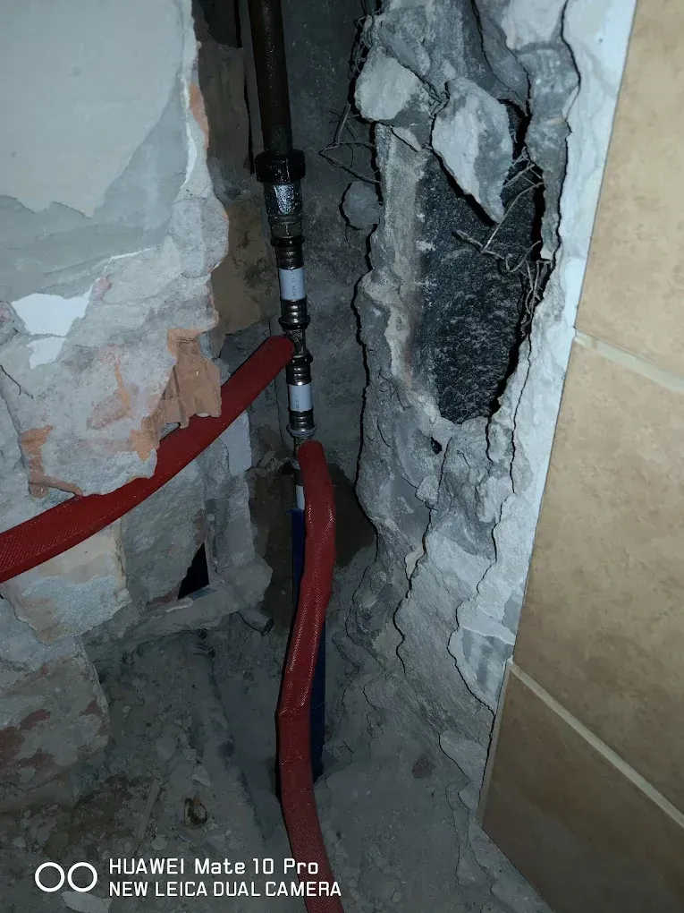 Egy kibontott falrészlet, amelyben a régi és új vízvezeték-hálózat találkozása látható (a 68-as kép távolabbi nézete). A nyers tégla és beton között egy régi fekete fémcső függőlegesen fut össze egy új, fehér műanyag csővel. Ebből a gerincből kék és piros szigetelésű (gégecsöves) ágak futnak lefelé és balra a padló irányába. A fal jobb oldalán érintetlen, világosbarna csempe látszik. A kép alján Huawei vízjel szerepel.