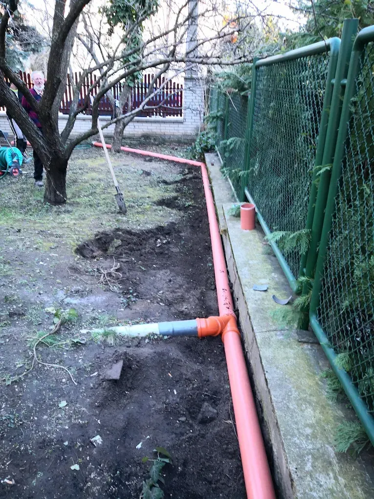 Szabadtéri jelenet egy kertben, ahol csatornázási vagy vízelvezetési munkálatok zajlanak. A földben egy frissen ásott árok fut egy zöld drótkerítés mentén, amelyben hosszú, narancssárga PVC lefolyócsöveket fektettek le. Egy Y-idomnál egy szürke cső is csatlakozik a narancssárga fővezetékhez. A háttérben fák, egy ásó, és egy fa mellett álló idős férfi látható.