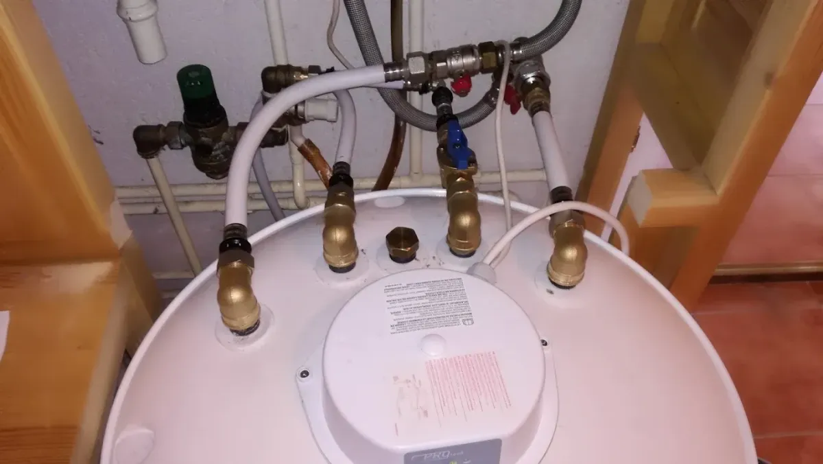 Egy fehér színű, hengeres elektromos vízmelegítő (bojler) teteje látható, PROtech feliratú műanyag burkolattal. A bojler tetejéből három sárgaréz könyökidom áll ki, amelyekhez fehér műanyag csövek csatlakoznak. A középső csőágon egy kék karos golyóscsap található. A bojler mögötti falon további gépészeti elemek, flexibilis fémcsövek, piros csapok és egy nyomáscsökkentő szelep látható felszerelve.