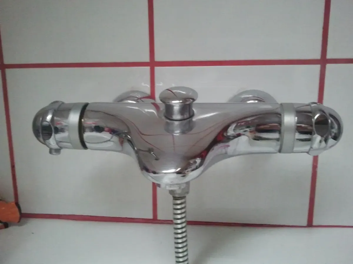 Közeli kép egy krómozott, fali kád- és zuhanycsaptelepről. A csaptelep két oldalán forgatható gombok vannak, középen pedig egy felhúzható zuhanyváltó gomb található. Alulról egy fém spirálos zuhanycső csatlakozik hozzá. A háttérben fehér négyzet alakú csempék láthatók, jellegzetes, vastag piros fugával.