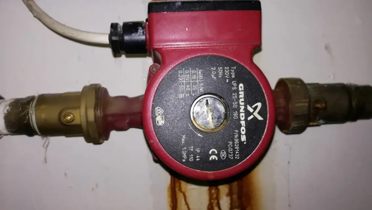 Közeli felvétel egy piros színű Grundfos fűtési keringető szivattyúról (típusa: UPS 25-50 180), amely egy rézcső-hálózathoz van csatlakoztatva. A szivattyú elején található a fekete adattábla a műszaki paraméterekkel és egy fém csavar középen. A szivattyúból egy fehér elektromos kábel vezet el felfelé. A háttérben lévő fehér falon a szivattyú alatt rozsdás, barna folyásnyomok láthatók.