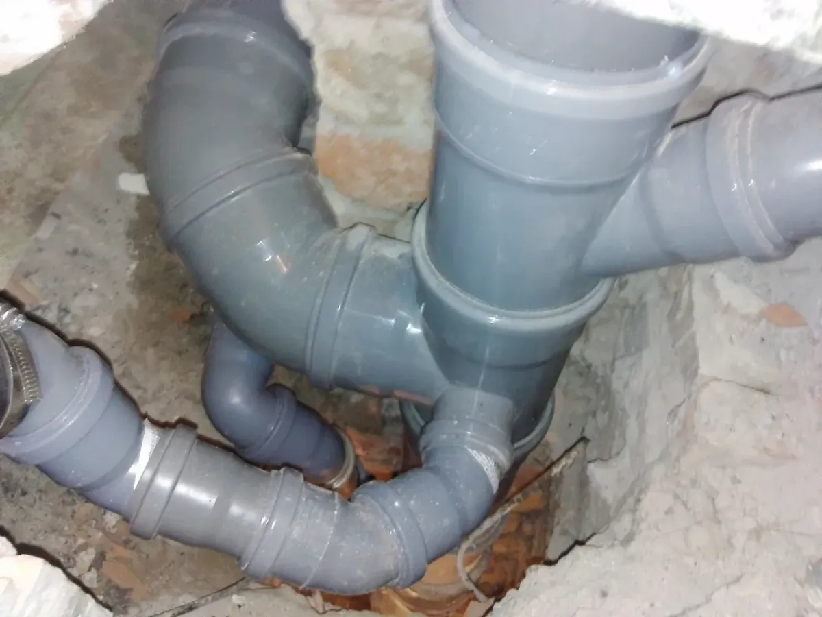 Falba vagy padlóba süllyesztett, vastag szürke PVC lefolyócsövek (csatornacsövek) bonyolult csomópontja látható egy kibontott nyílásban. A fővezetékhez több Y-idommal és könyökkel csatlakoznak vékonyabb ágak. Bal oldalon az egyik vékonyabb csövet egy fém bilincs rögzíti. A csövek körül nyers tégla, beton és törmelék látható.