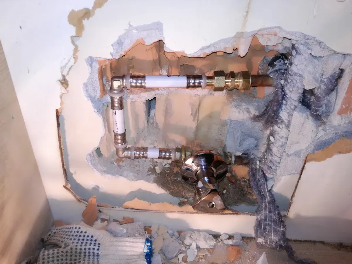 Egy falba vájt szabálytalan nyílás, amelyben vízvezeték-szerelési munkálatok láthatók. Két vízszintes, fehér ötrétegű csődarabot toldottak be sárgaréz présidomokkal a meglévő hálózatba. Alattuk egy krómozott, háromszögletű elzárócsap (fogantyú) áll ki a falból. A bontott üregben tégla, vakolat és egy rongy is látszik, a kép alján pedig egy munkáskesztyű hever a törmelék között.