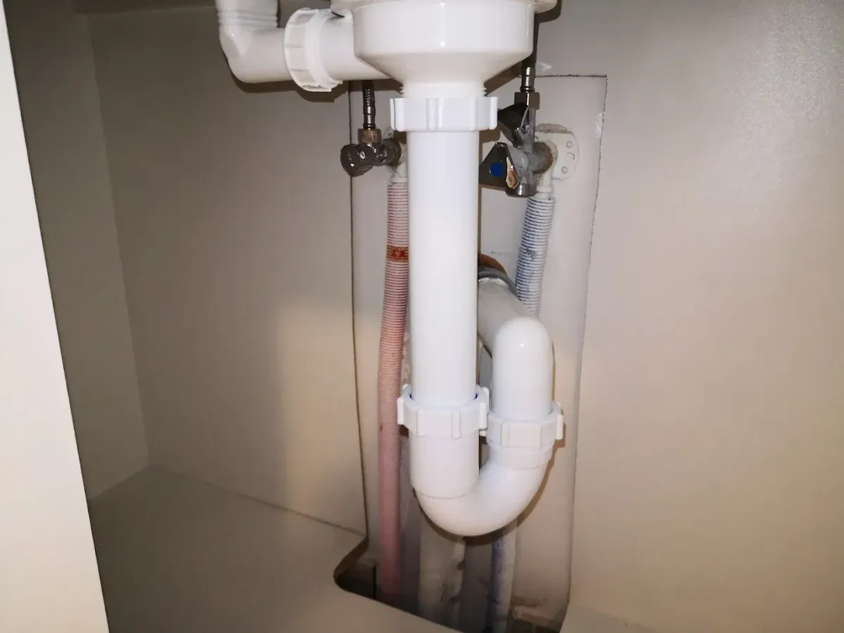 Mosdó alatti, U-alakú fehér műanyag szifon és lefolyócső látható egy szekrényben. A háttérben egy falbontáson keresztül látszanak a sarokszelepek, a piros/kék szigetelőcsövek és a flexibilis csatlakozások.