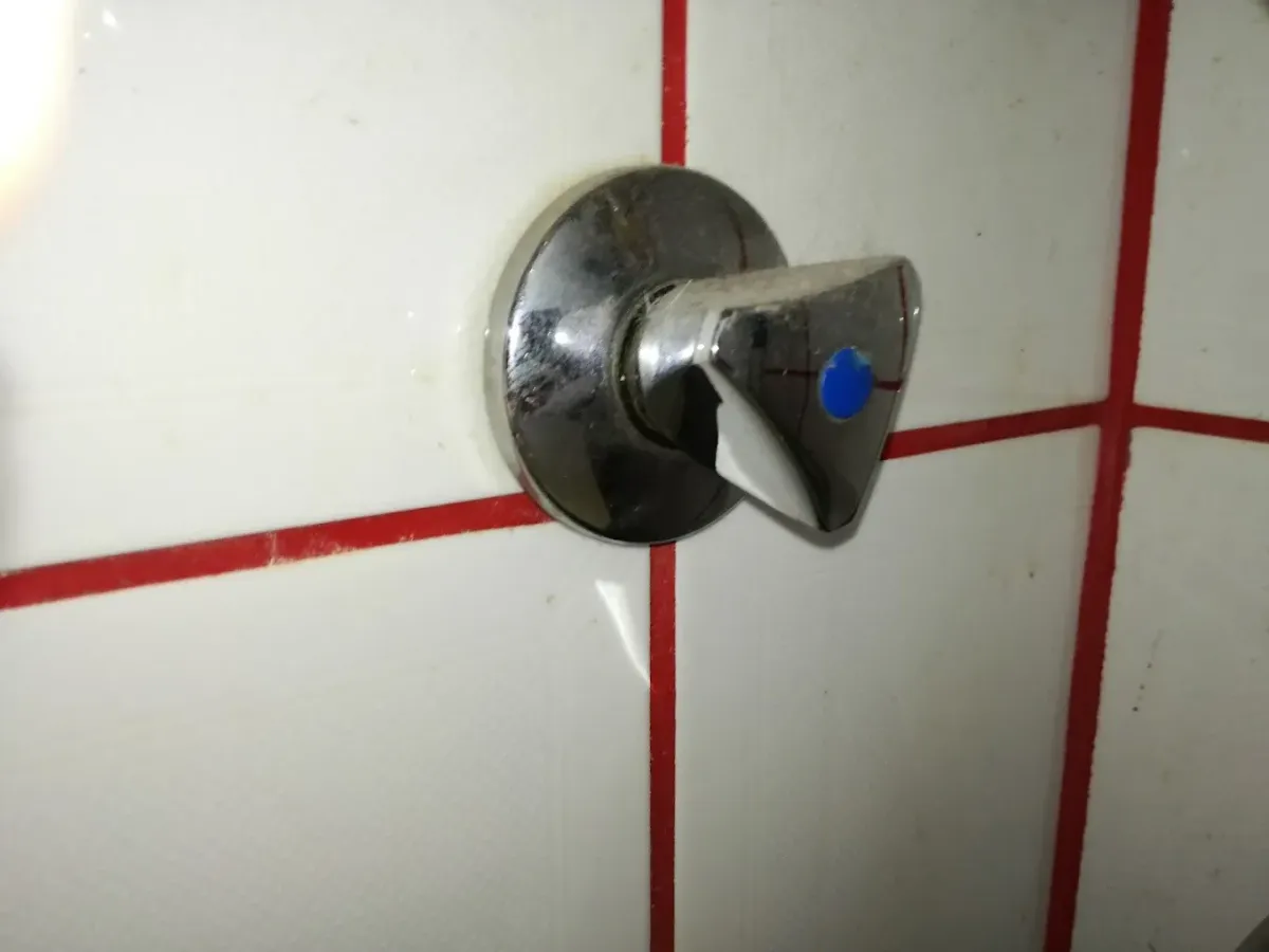 Nagyon közeli kép egy falra szerelt, háromszög formájú, krómozott elzárócsap-fogantyúról. A fogantyú közepén egy kis kék pötty jelzi a hideg vizet. A csap kissé vízköves és poros, a mögötte lévő falat fehér csempe borítja, feltűnő és vastag piros fugával.