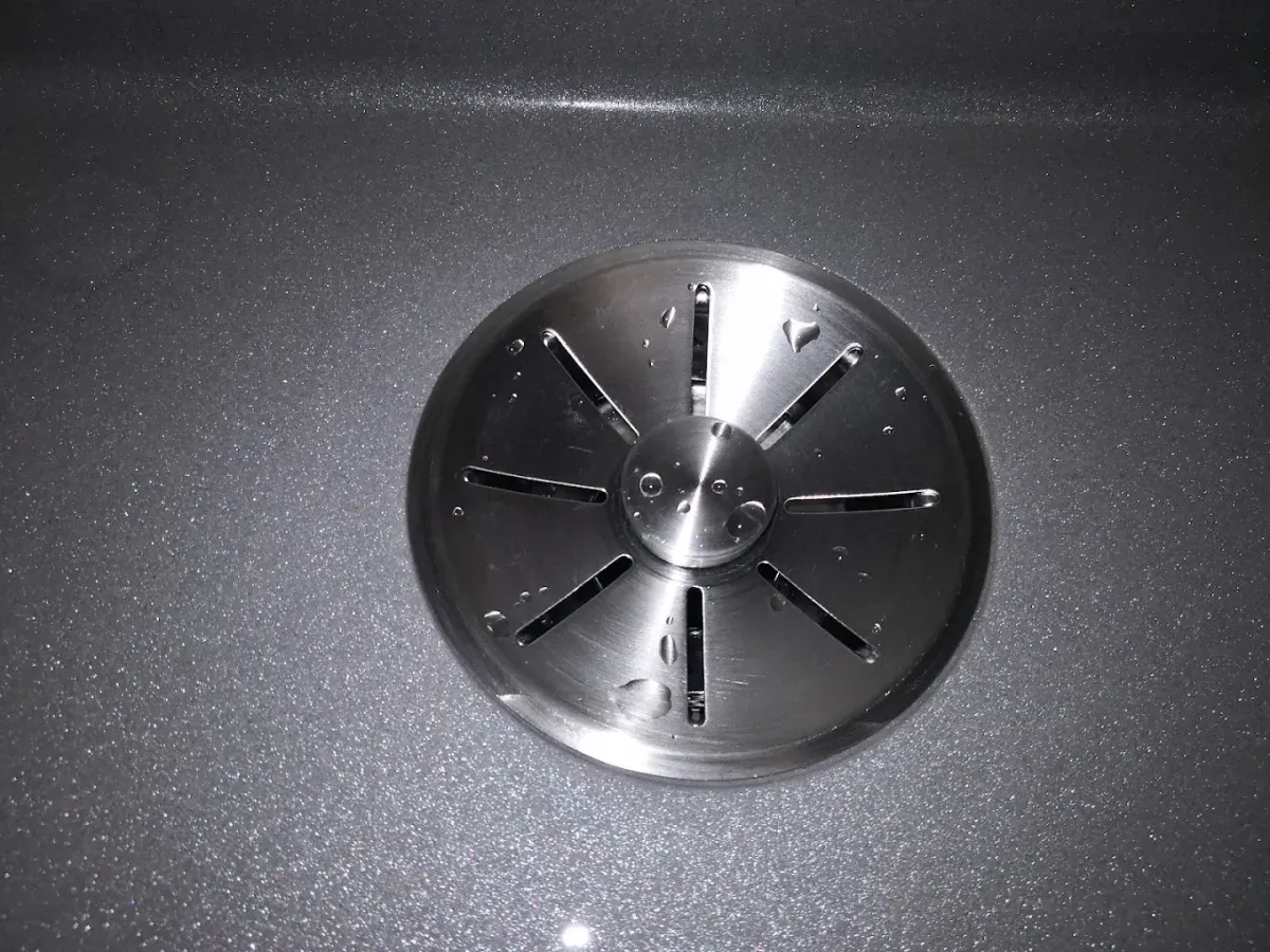 Nagyon közeli, felülnézeti kép egy sötétszürke, csillogó felületű (valószínűleg gránit vagy kompozit) konyhai mosogatótálca lefolyójáról. A lefolyót egy kerek, szálcsiszolt rozsdamentes acél szűrő vagy dugó fedi, amelyen sugárirányban elhelyezkedő rések és egy középső fém gomb található. A felületen néhány apró vízcsepp is megcsillan.