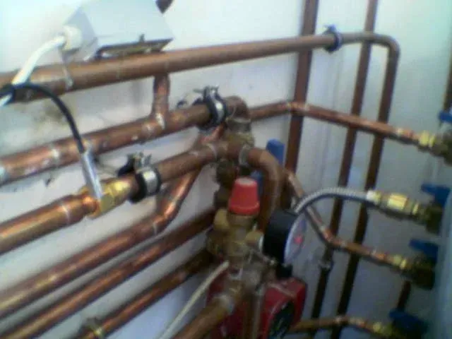 Gépészeti helyiség vagy kazánház falán futó, bonyolult vörösréz csőhálózat közelképe. A csövek sárgaréz idomokkal és bilincsekkel vannak rögzítve. Középen egy piros kupakos biztonsági szelep vagy légtelenítő, valamint egy nyomásmérő óra (manométer) látható. Kék karos golyóscsapok és elektromos vezetékek, köztük egy fehér kötődoboz is megfigyelhetők a rendszeren.