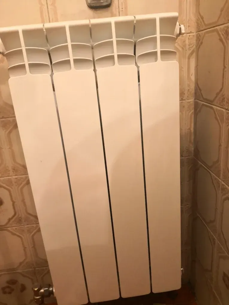 Egy négy tagból álló, függőleges kialakítású, lapos paneles fehér radiátor (fűtőtest) látható. A radiátor egy régebbi stílusú, barna és bézs színű, geometrikus mintás csempével burkolott falra van felszerelve. A radiátor bal alsó sarkában egy apró sárgaréz szelep és csőcsatlakozás is megfigyelhető.