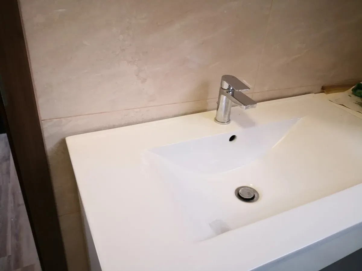 Egy modern, szögletes kialakítású, széles peremű fehér kerámia mosdókagyló látható felülről fotózva, egy pulton. A mosdón egy elegáns, lapos formájú, krómozott egykaros csaptelep van. A medence alján krómozott lefolyódugó látszik. A hátteret nagyméretű, világos bézs, homokszínű falicsempék alkotják.