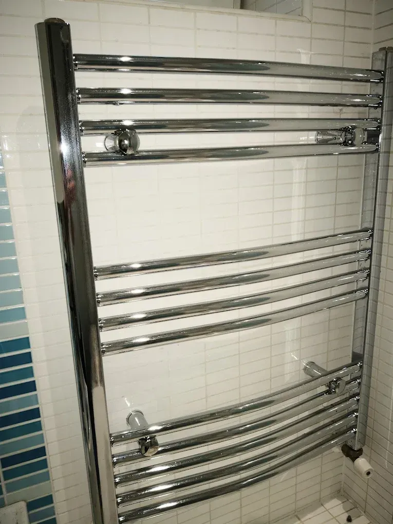 Egy fürdőszobai, falra szerelt, fényes krómozott törölközőszárító radiátor látható. A radiátor csövei vízszintesen, enyhe ívben hajlítva futnak. A falat fehér, apró négyzetes csempe borítja, amelynek bal oldalán egy függőleges, kék árnyalatú üvegmozaik díszítőcsík húzódik. A radiátor alsó részén szelepek és fűtéscső-bekötési pontok láthatók.