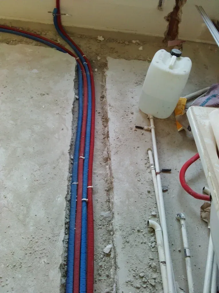 Egy felújítás alatt álló helyiség nyers betonpadlója látható. A padlóba vágott horonyban három szigetelt víz- vagy fűtéscső fut egymás mellett (két kék és egy piros). A csatornától jobbra a betonon szabadon futó fehér műanyag vízvezetékcsövek, egy nagy fehér műanyag kanna, és némi építési hulladék hever.