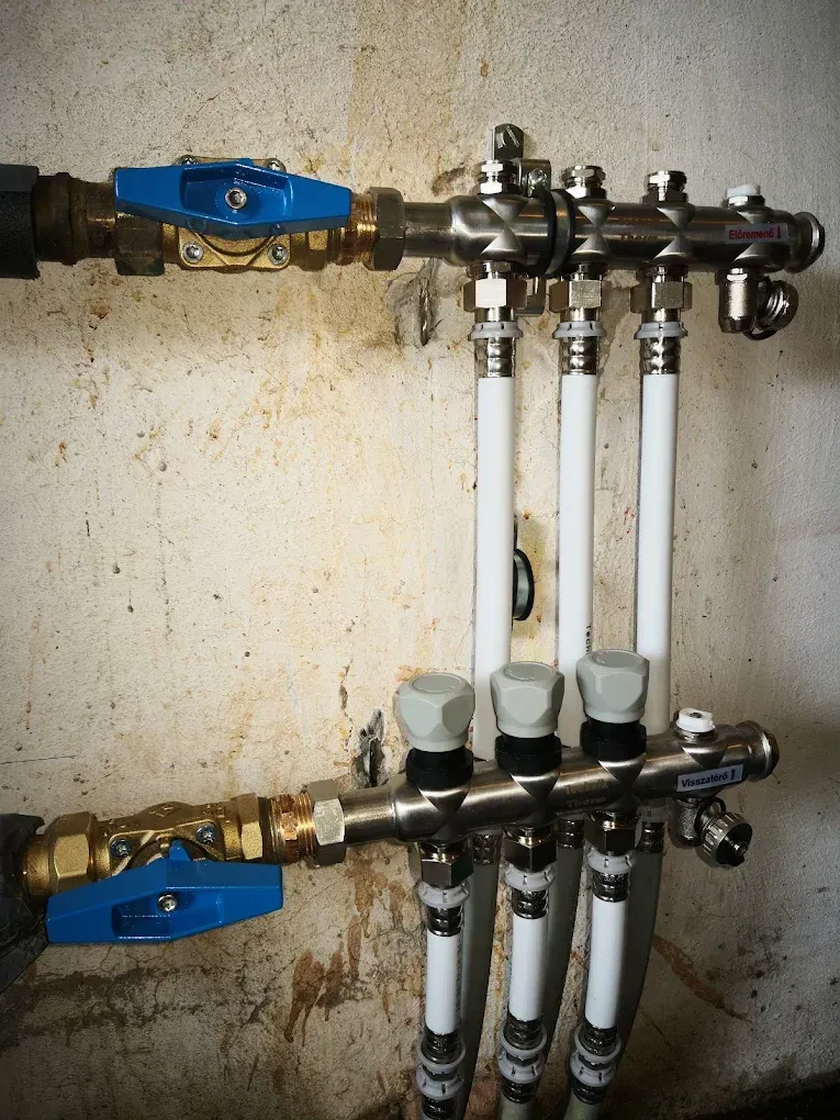 Nyers, vakolatlan falra szerelt rozsdamentes acél fűtési osztó-gyűjtő (elosztóblokk) látható. Két vízszintes csőoszlopból áll, mindkettő bal végén egy-egy vastag, kék karos golyóscsap található. A felső és alsó blokkhoz függőlegesen fehér ötrétegű csövek csatlakoznak fém csavarzatokkal. Az alsó tagon világosszürke műanyag védőkupakok vannak a szelepeken, a csöveken pedig kék és piros feliratok látszanak.