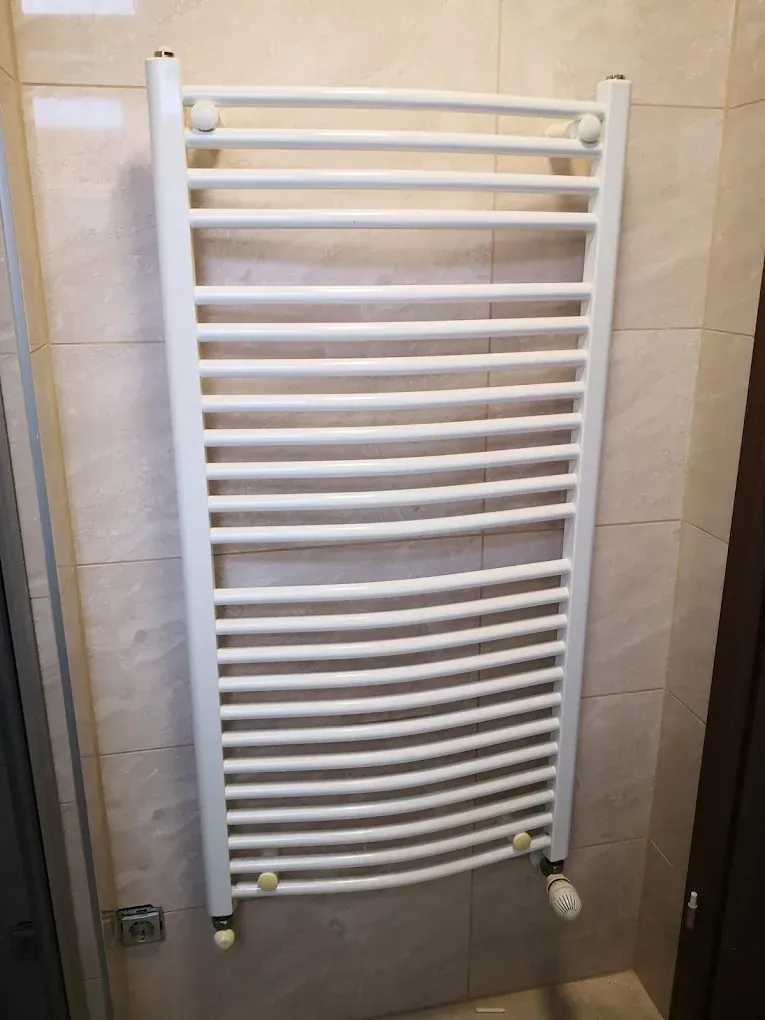 Egy fehér színű, csöves, falra szerelt fürdőszobai törölközőszárító radiátor látható. A falat nagyméretű, világos bézs színű, márványos mintázatú csempe borítja. A radiátor jobb alsó sarkában egy fehér termofej (hőfokszabályozó szelep) található. Bal oldalon lent egy fedél nélküli, falba süllyesztett elektromos kötődoboz vagy aljzat látszik.