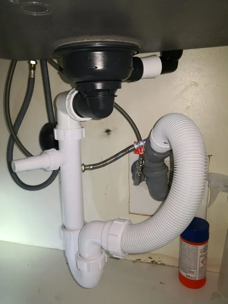 Konyhai vagy fürdőszobai mosogató alatti gépészet. Felül a fekete lefolyócsonk látható, amihez fehér műanyag csövek és egy U-alakú szifon csatlakozik. Jobb oldalon egy hurokba hajlított, vastag, bordázott fehér gégecső fut. A háttérben flexibilis fém vízbekötő csövek és egy falból kiálló sarokszelep található, jobb oldalt lent pedig egy narancssárga-kék flakon (tisztítószer) áll.