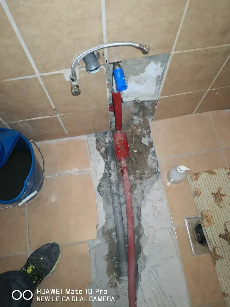 Csőtörés javítása vagy fürdőszoba felújítása során feltört padló és fal látható. A padlócsempék között a nyers betonig bontott sávban egy piros szigeteléssel ellátott cső fut a fal felé. A falból megbontott csempék mögül egy kék védőkupakos kiállás, egy flexibilis cső és egy szürke PVC lefolyócsonk áll ki. A kép bal szélén egy kék felmosóvödör, alul egy cipő orra látszik, jobbra pedig egy szappanadagoló és egy tengeri mintás kilépőszőnyeg található. A kép alján "HUAWEI Mate 10 Pro" vízjel olvasható.