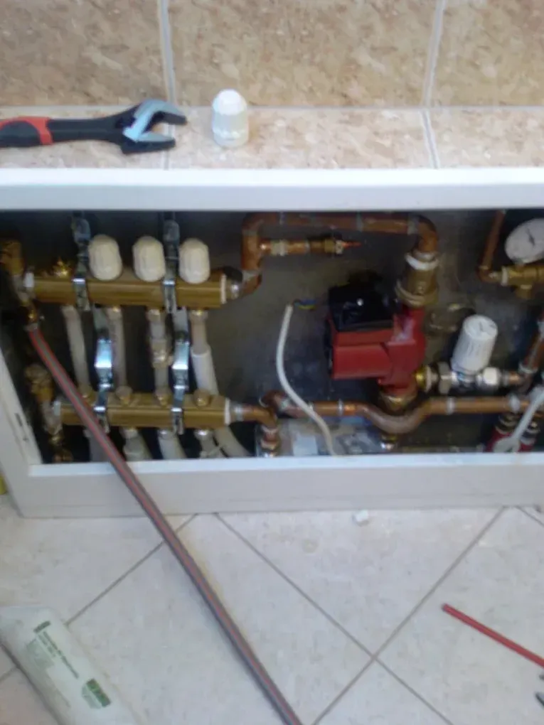 Egy falba süllyesztett fehér fém dobozban (osztó-gyűjtő szekrényben) lévő fűtési rendszer szerelvényei láthatók. Bal oldalon egy sárgaréz osztó-gyűjtő blokk található három fehér termofejjel és rácsatlakozó műanyag csövekkel. Jobb oldalon vörösréz csövek, szelepek, egy nyomásmérő óra és egy piros színű keringető szivattyú kapott helyet. A szekrény alatti padlócsempén egy hosszú, színes szigetelésű vezeték, felül a párkányon pedig egy fekete-piros villáskulcs és egy fehér termofej hever.