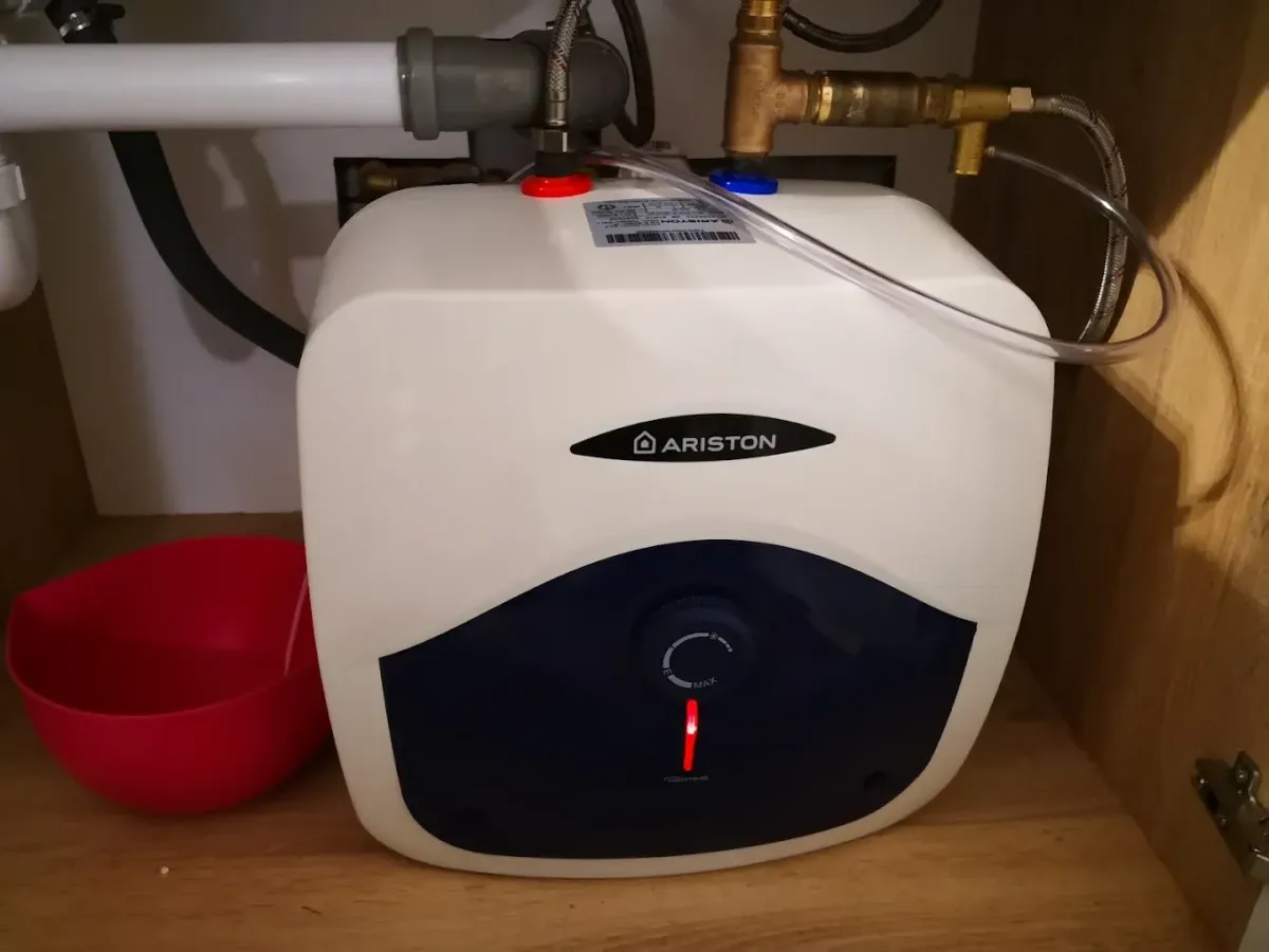 Mosogató vagy mosdó alatti szekrényben felszerelt, kisméretű Ariston elektromos vízmelegítő (bojler) látható. A készülék fehér színű, alul sötétkék panellel, amelyen egy tekerőgomb és egy világító piros visszajelző lámpa (fűtésjelző) található. A bojler tetejéhez piros és kék gyűrűvel jelölt vízcsövek és egy sárgaréz biztonsági szelep csatlakozik egy átlátszó műanyag csővel. Bal oldalon egy szürke szifon részlete és egy piros műanyag tál látszik a fa erezetű szekrény alján.