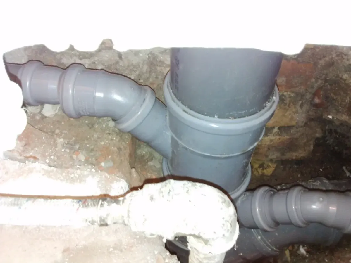 Kibontott téglafal és beton padlózat részlete, amelyben vastag szürke PVC lefolyócsövek (csatornacsövek) láthatók. A fővezetékhez egy Y-idommal egy másik cső csatlakozik, alatta pedig egy kisebb átmérőjű lefolyócső is be van kötve. Az előtérben egy fehér, gipsszel vagy szigeteléssel bevont csatlakozó is feltűnik a törmelék között.
