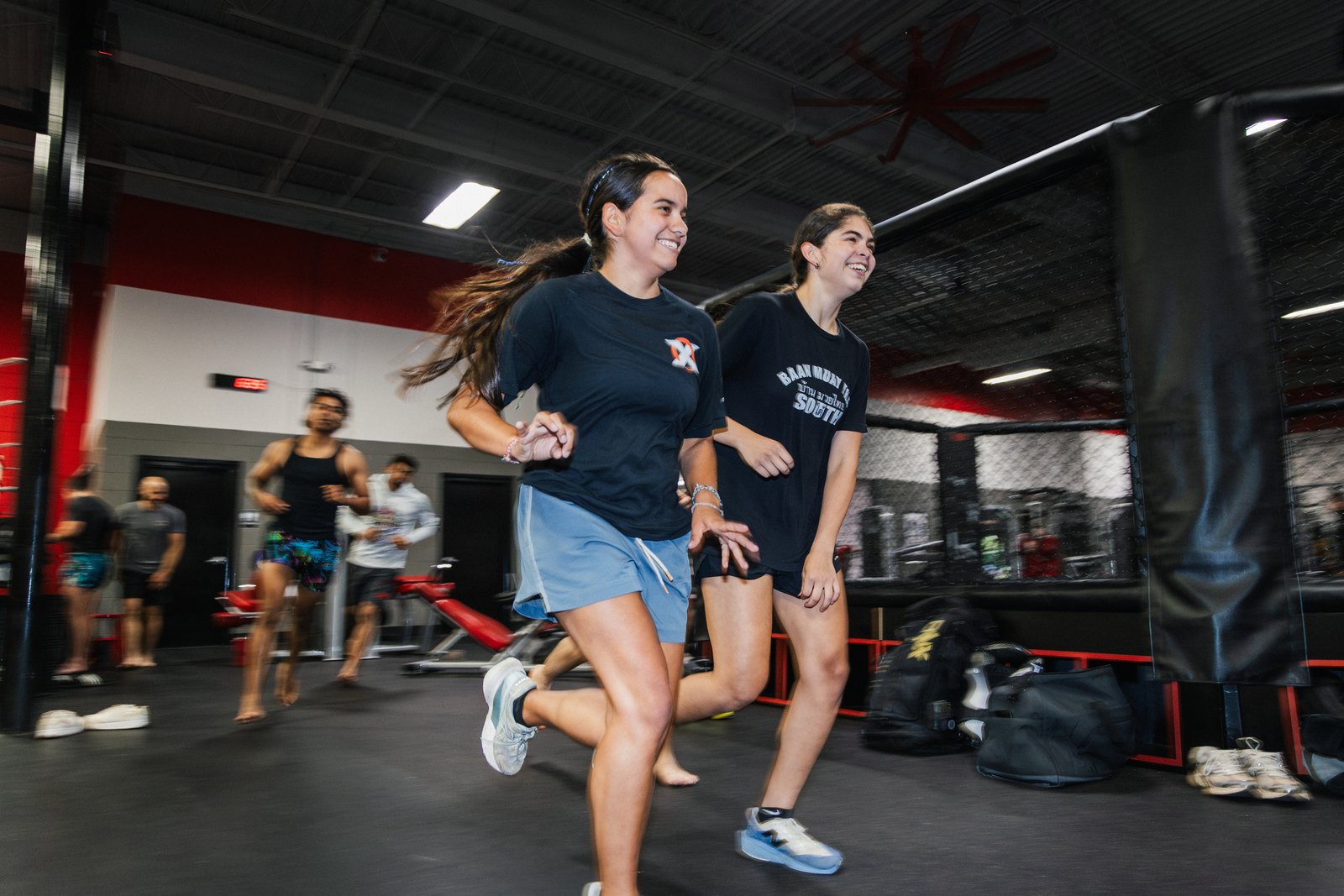 Two women running laps together on the gym floor at X-Zen Sports, smiling, with the MMA cage and weight area behind them