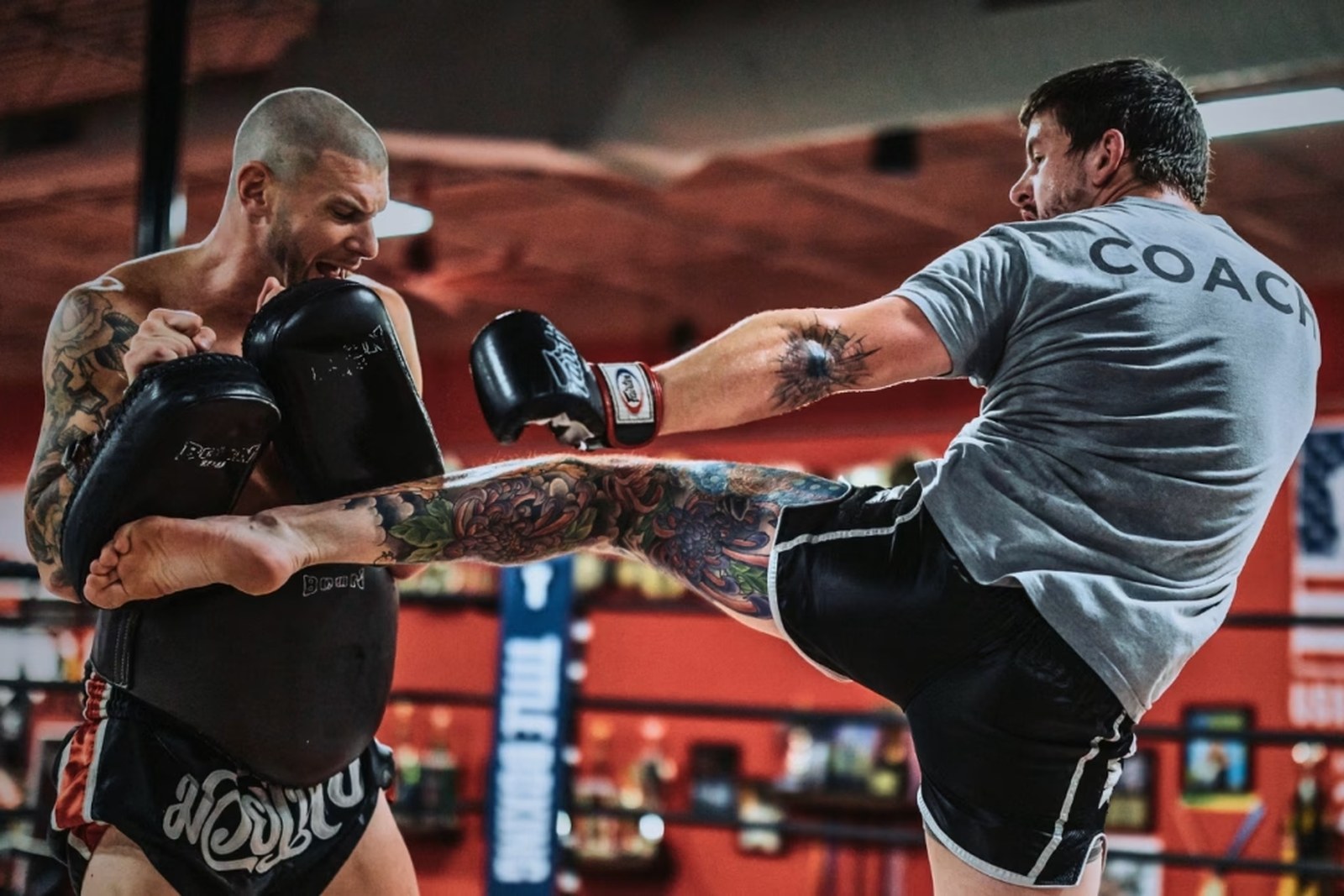 A Muay Thai coach at X-Zen Sports landing a high kick on the pads during partner drills, with the boxing ring behind them