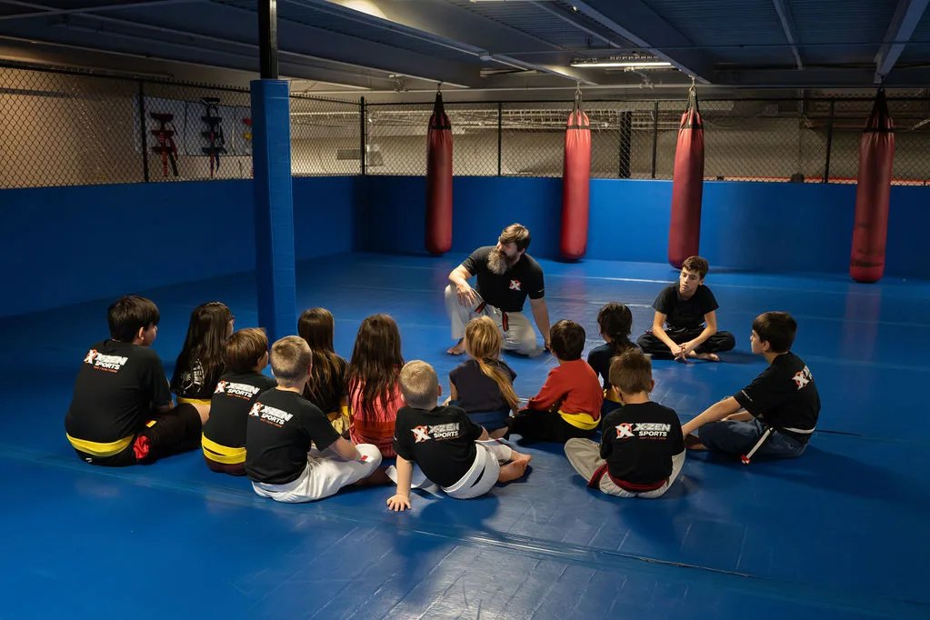 Coach Cody Freeland talks with a circle of young students on the mats at X-Zen Sports.
