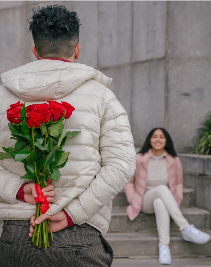 smiling woman sitting on steps looking at a man holding red roses behind his back